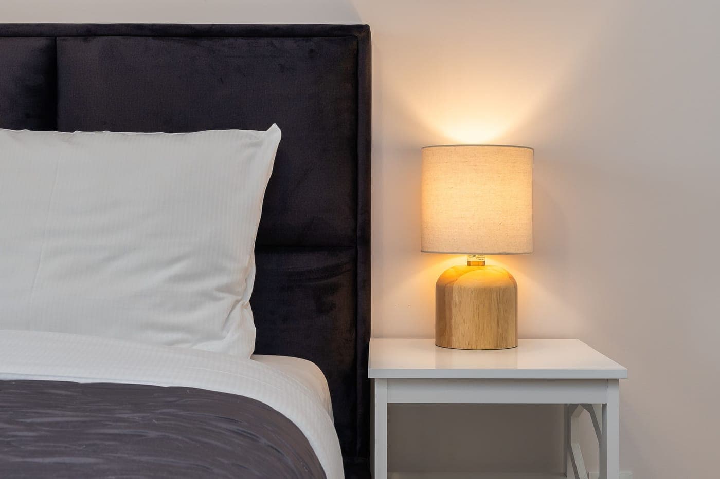 A warmly lit lamp on a white nightstand beside a bed